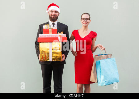 L'homme d'affaires barbu joyeux dans Red Hat et femme d'affaires en robe rouge et verres, tenant des cadeaux de Noël et les paquets colorés et à la recherche au camer Banque D'Images