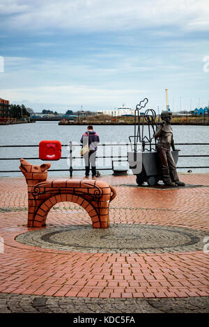 Un homme se tient debout en regardant un téléphone portable au-dessus du bassin de Roath un jour humide de printemps, la sculpture Beastie Bench and Coal Miner au premier plan, Cardiff Bay, pays de Galles, Royaume-Uni Banque D'Images