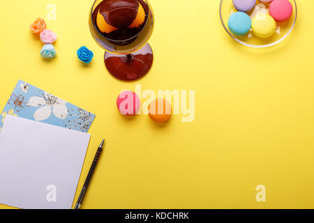 Macarons colorés et de verre sur le tableau bleu avec stylo et carte postale sur fond jaune Banque D'Images