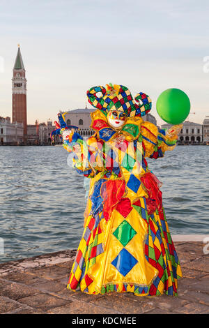 Carnaval de Venise 2017, Venise, Italie. Arlequin femme de couleur vive jester posant au coucher du soleil avec la lagune et Campanile derrière elle Banque D'Images