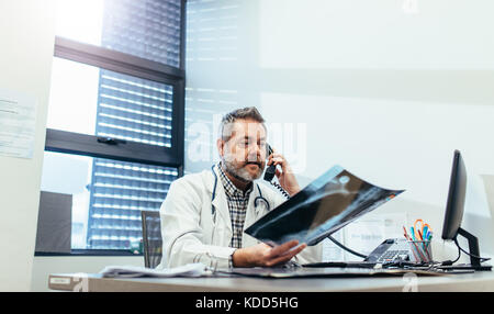 Le radiologue contrôle d'un rapport d'analyse médicale et parlant au téléphone. Mature doctor examining x-ray et à l'aide de téléphone dans sa clinique. Banque D'Images