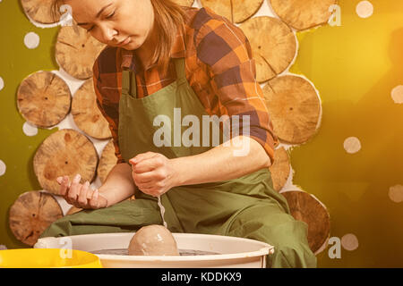 Une jeune femme en jeans et chemise à carreaux verse de l'eau sur l'argile pour l'adoucir et prendre un bol sur un tour de potier dans un cadre lumineux, belle déco atelier Banque D'Images