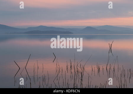 Un lac au crépuscule, avec de belles et chaudes dans le ciel et l'eau, réflexions sur les montagnes et les collines lointaines, les branches en premier plan Banque D'Images