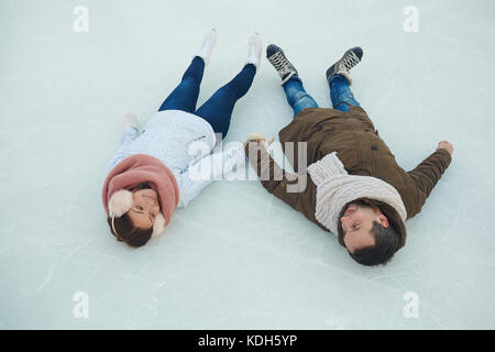 Amoureux heureux dans winterwear allongé sur une patinoire et tenue par les mains Banque D'Images