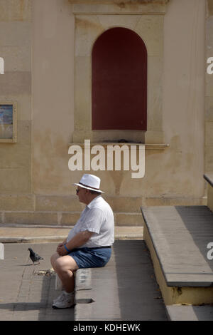 Un homme âgé portant des shorts sur mesure et un traditionnel chapeau panama assis sur des marches de pierre à l'extérieur d'une église à kerkira, Corfou, Grèce dans le soleil Banque D'Images