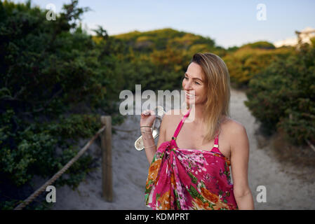 Jolie femme mince transportant ses chaussures aux pieds nus comme elle marche le long d'une plage de sable fin sourire joyeusement à chemin l'appareil photo en vacances Banque D'Images