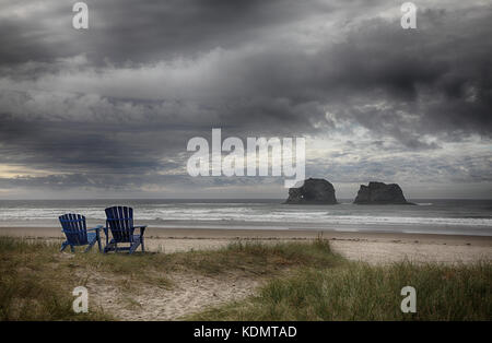 Un romantique oceanscape avec moody sky dont deux chaises Adirondack auxquels font face à la mer des formations de roche pile à Rockaway Beach, Oregon. Banque D'Images