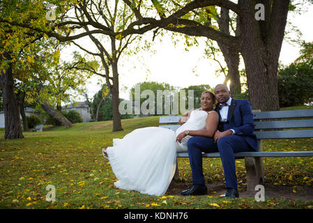 Jeune mariée et se toilettent assis sur un banc de parc au coucher du soleil dans le décor de l'automne Banque D'Images