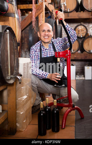 Cheerful man wearing apron en utilisant un appareil de bouchage bouteille wine house Banque D'Images