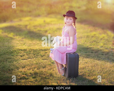 Cute little girl in a hat est assise sur une valise Banque D'Images