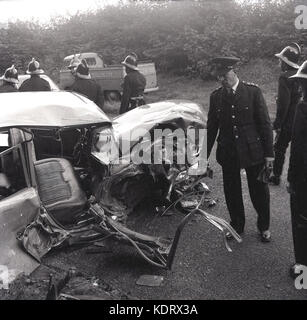 1960, España, poiice et pompiers avec des casques sur les lieux d'un accident de la route en milieu rural et une forte smashed up motorcar allongé sur la route. Banque D'Images