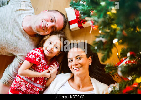 Les jeunes parents avec leur petite fille en vertu de l'arbre de Noël se prélassant parmi les cadeaux. Banque D'Images