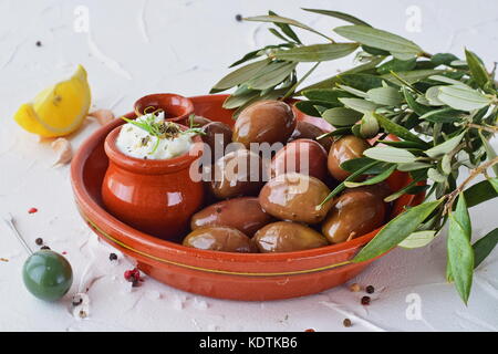 Les olives dans un bol en céramique marron traditionnel, avec sauce tzatziki, citron, ail, branche d'olivier et l'huile d'olive sur un fond blanc. Banque D'Images