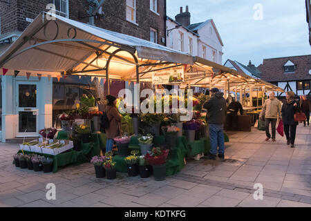 Soir dans le centre de York avec des gens qui marchent par allumé en pagaille les étals de marché commerçant et par son comité permanent de décrochage de fleurs en premier plan - England, UK. Banque D'Images