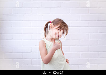 Little girl with Thumbs up. Attitude positive. Jolie petite fille blonde enfant giving Thumbs up et des clins d'œil viewer. Cute kid contre bric blanc Banque D'Images