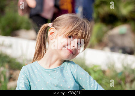 Belle jeune femme enfant. cheveux blonds fille avec sourire effronté et les yeux bruns portant un top vert souriant à la visionneuse sur une journée ensoleillée. Banque D'Images