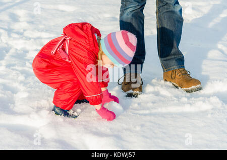 Cute toddler kid jouer dans la neige. Petite fille enfant accroupi faire une boule de neige à côté de mâle adulte, jambes et pieds. L'hiver de vie actif. Banque D'Images