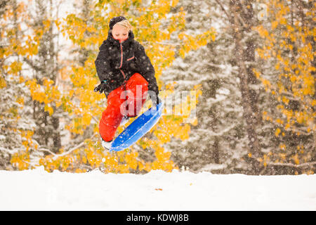 Un garçon saute sur son disque en faisant du traîneau à neige pendant une chute de neige à la fin de l'automne à Redwood Meadows, Alberta, Canada. Banque D'Images