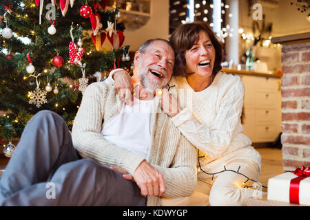 Couple assis sur le sol en face de l'arbre de Noël lumineux à l'intérieur de leur maison, parler et rire. homme empêtré dans la chaîne de lumières. Banque D'Images