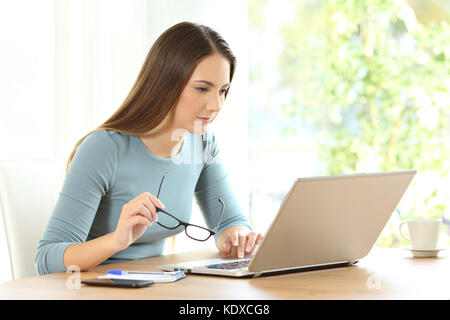 Femme sérieuse holding eyeglasses lecture le contenu en ligne un ordinateur portable sur une table à la maison Banque D'Images