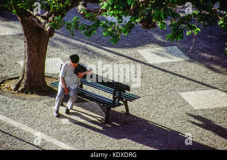 Un homme âgé est assis seul sur un banc de parc à Angra, Hiroismo l'île de Terceira, aux Açores. La route et le trottoir sont incrustés cobbletone avec dessins et modèles. Banque D'Images