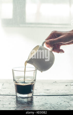 Personne versant le lait des jug dans un verre de café noir sur la table en bois Banque D'Images