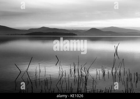 Un lac au crépuscule, avec des tons doux, beau dans le ciel et l'eau, réflexions sur les montagnes et les collines lointaines, les branches en premier plan Banque D'Images