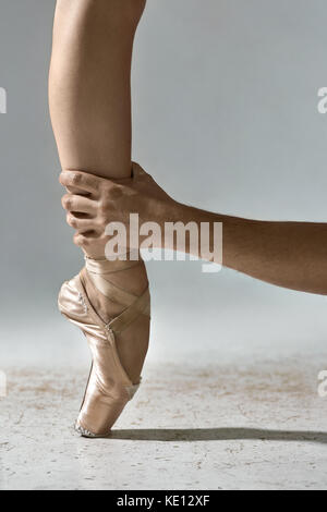 La main de l'homme tient la jambe de la ballerine sur fond gris en studio. girl porte des chaussures beige pointe et se tient sur la toe. libre verticale. Banque D'Images