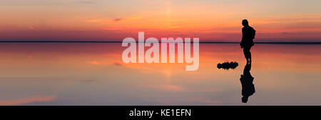 Silhouette de l'homme seul à la dynamique vers le coucher du soleil reflétée dans les eaux peu profondes du lac solt taille de bannière. Banque D'Images
