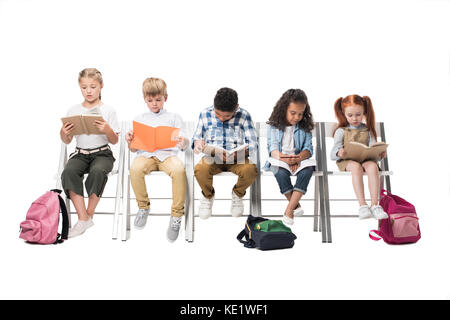 La lecture de livres pour enfants multiethnique tout en étant assis sur des chaises isolated on white Banque D'Images