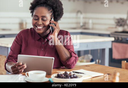 Souriante jeune femme africaine l'utilisation d'un cellulaire et à l'aide d'une tablette numérique tout en étant assis à une table de travail à domicile Banque D'Images