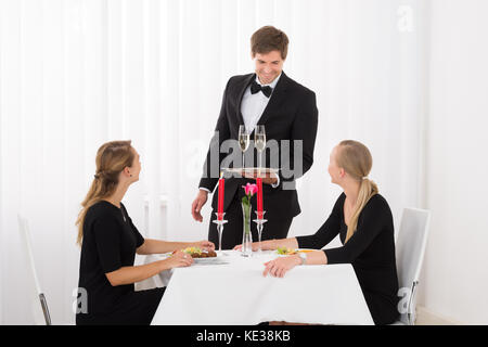 Happy Waiter Serving verre de champagne pour amies au restaurant avec de la nourriture sur la table Banque D'Images