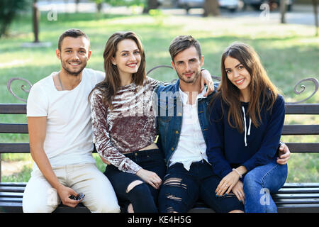 Quatre femmes et hommes assis sur un banc à l'embrasser. heureux jeune élégant friends posing on bench outdoors, smiling at camera et en bonne santé. Banque D'Images