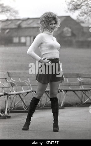 Années 1960, historiques, femme blonde bien roulée avec de grands cheveux ondulés posant dehors dans une mini jupe noire et des bottes aux genoux. Banque D'Images