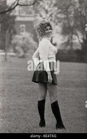 Années 1960, historiques, femme blonde bien roulée avec de grands cheveux ondulés posant dehors dans une mini jupe noire et des bottes aux genoux. Banque D'Images