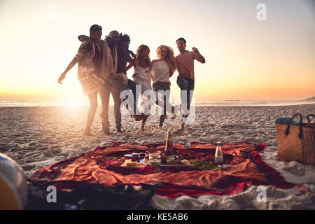 De jeunes amis ludiques se mettent à pied, en pique-nique sur la plage ensoleillée au coucher du soleil Banque D'Images