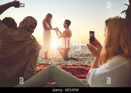 Jeune homme proposant à la femme sur la plage ensoleillée d'été avec amis Banque D'Images