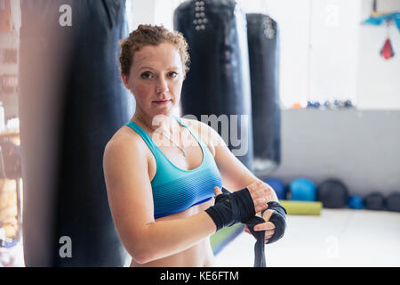 Portrait confiante, robuste caleçons femme enveloppant les poignets à côté de sacs de poinçonnage dans la salle de gym Banque D'Images