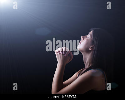 Gros plan d'une femme fidèle priant sous une lumière céleste ou divine, mains jointes en prière, la tête et les yeux fermés dans l'inspiration religieuse de la prière. Banque D'Images