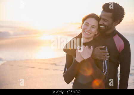 Couple multiethnique souriant et affectueux en costume humide sur une plage ensoleillée au coucher du soleil Banque D'Images