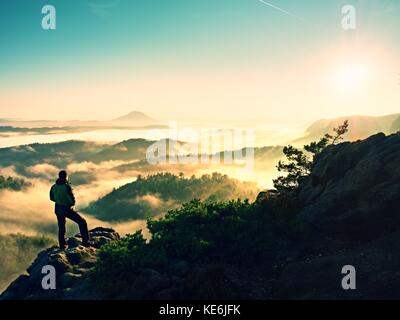Silhouette homme haut escalade sur falaise. randonneur est montée jusqu'au pic de profiter de la vue. l'homme veille sur misty et matin brumeux en vallée de soleil du matin. Banque D'Images