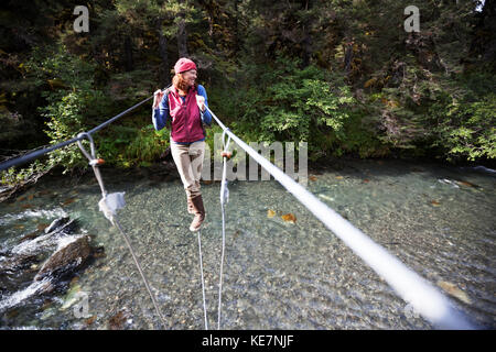 Une femme marche sur un câble sur un pont suspendu au-dessus de la rivière ; Alaska, États-Unis d'Amérique Banque D'Images