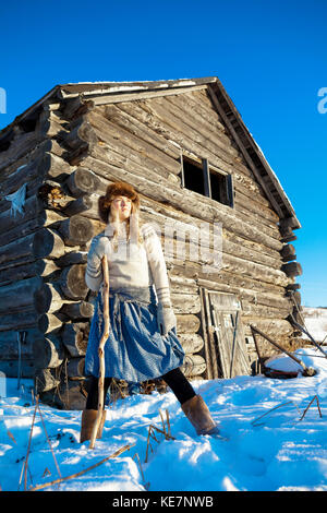 Une fille avec un chapeau de fourrure se trouve dans la neige avec un bâton de marche et une cabane en arrière-plan ; Homer, Alaska, United States of America Banque D'Images