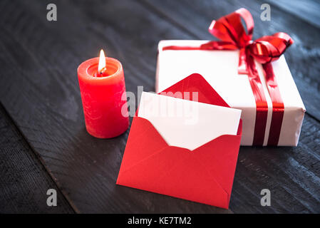 Enveloppe rouge vide s'appuya contre un cadeau emballé avec style, attachés avec du ruban rouge et Bow, près d'une bougie allumée sur une vieille table en bois. Banque D'Images