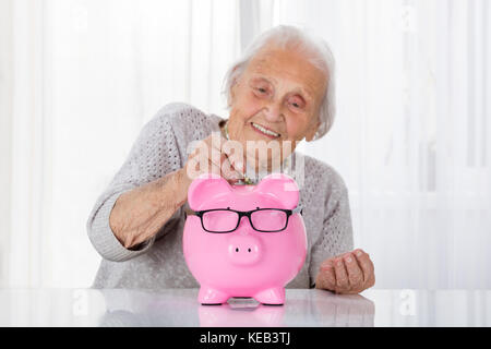 Happy Senior Woman Inserting Coin In Piggybank On Table Banque D'Images