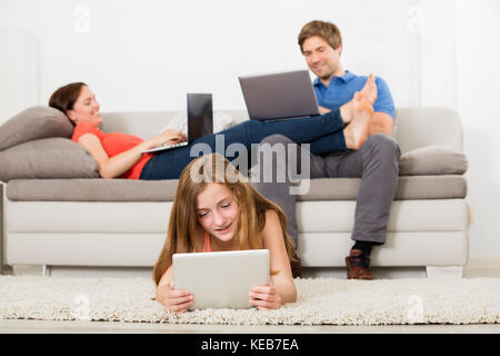 Girl Lying On Carpet Using Digital Tablet in front of Parent Working On Laptop Banque D'Images