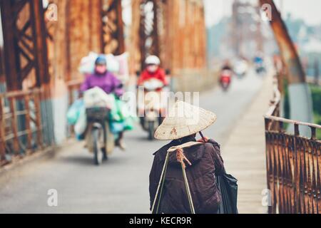 Vendeur de rue vietnamiens mène beaucoup de choses à travers le pont long bien à Hanoï, au Vietnam. selective focus sur la coiffe traditionnelle. Banque D'Images