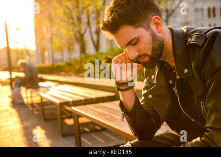 Portrait d'un beau jeune homme hipster assis sur un banc dans le coucher du soleil et de parler sur son téléphone Banque D'Images