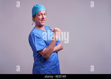 Un médecin de sexe masculin d'âge moyen se tenant en toute confiance dans un gommage bleu et se pointant vers lui-même Banque D'Images