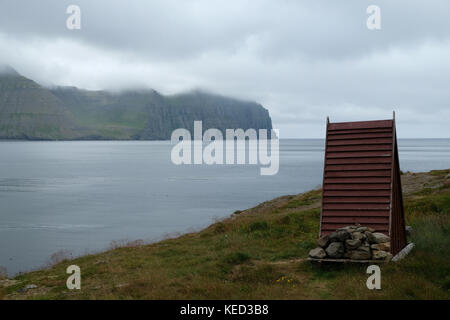 Old outhouse rouge triangulaires en bois donnant sur l'océan et les montagnes ancrée par des pierres contre le vent dans la réserve naturelle de hornstrandir, Islande Banque D'Images